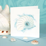Quilled Abstract Nautilus Greeting Card standing up in front of an ocean background, surrounded by seashells.