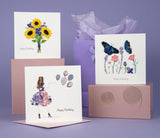 Quilled Birthday Flowers & Blue Butterflies Greeting Card sitting on top of a Quilling Card Storage Box, in front of a purple gift back and background, next to Quilled Birthday Sunflower Bouquet Greeting Card and Quilled Fashion Birthday Girl Card.