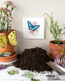 Quilled Blue Butterfly & Pink Flowers Greeting Card on white brick wall above pile of soil with gradening shovel on table with terra cotta pots with plants in them