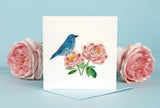 Quilled Bluebird & Peonies Greeting Card standing up in front of a light blue background with pink flowers behind it.