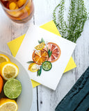 Quilled Citrus Art Greeting Card with yellow envelope next to plate of limes and lemons, with glass of iced tea on white wooden background