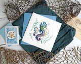 Quilled Colorful Seahorse Greeting Card laying on a dark blue napkin next to Quilled Sand Dollar Gift Enclosure Mini Card, surrounded by nets and a postcard.