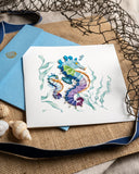 Quilled Colorful Seahorse Greeting Card laying on top of a knitted backdrop, next to a navy ribbon, a net, and seashells.
