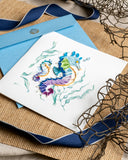 Quilled Colorful Seahorse Greeting Card laying on top of a knitted backdrop, next to a navy ribbon, a net, and seashells.