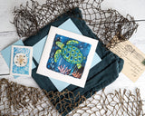 Quilled Decorative Sea Turtle Greeting Card with light blue envelope on dark teal envelope next to netting, postcard, and quilled sand dollar miniature gift enclosure on white wooden background
