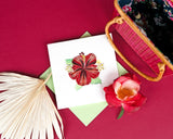 Quilled Hibiscus greeting card laying flat on maroon background surrounded by a palm leaf, a  flower, and a Pink Hand-woven Bamboo Bag.