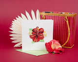 Quilled Hibiscus greeting card standing up in front of a maroon background surrounded by a palm leaf, a flower, and a Pink Hand-woven Bamboo Bag.