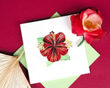 Quilled Hibiscus greeting card laying flat on maroon background surrounded by a palm leaf and a red flower.