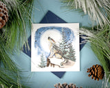 Quilled Howling Wolf Greeting Card laying flat on a light blue backdrop, beneath pine needles and pinecones.
