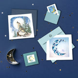 Quilled Howling Wolf Greeting Card laying on a dark blue background, covered in glistening stars, besides a Quilled Crescent Moon Greeting Card, Quilled Moon & Stars Gift Enclosure Mini Card, and Quilled Moon Sticky Note Pad Cover.