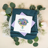 Quilled Hydrangea Bouquet Birthday Card with lilac envelope on dark blue napkin and plate surrounded by eucalyptus and baby's breath, on top of white lace doiley
