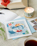 Quilled Koi Fish Pond Greeting Card laying on a linen cloth next to candles, stamps, a teacup, and open book with a flower.