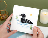 Quilled Loon greeting card being held in two hands in front of dark green background with candle