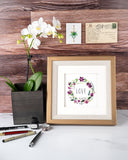 Quilled LOVE Floral Wreath Greeting Card in golden frame next to white orchid in front of wooden wall with postcard and mini hanging plant gift enclosure on white marble table with pens