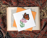 Quilled Monarch Milkweed Butterfly Greeting Card sitting on an open book laying over a patterned scarf.