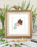 Quilled Monarch Milkweed Butterfly Greeting Card in a Golden Square Artist Series Frame sitting on a wooden surface besides pink flower vases and dry flowers.