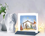 Quilled Nativity Scene Christmas Card standing with navy. blue envelope next to angel statue on white counter with bokeh lights behind and white wall