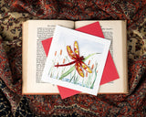 Quilled Red Dragonfly & Cattails Greeting Card with red envelope on top of open book on top of paisley tapestry