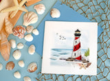 Quilled Red & White Lighthouse Greeting Card laying down sitting on top of a rope net, next to a wooden box full of seashells over a light blue background.
