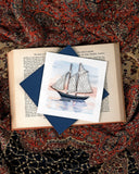 Quilled Schooner at Sunset Greeting Card laying on an open book on a patterned scarf.