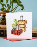 quilled vintage luggage greeting card standing up with red envelope in front of books and plant on light blue background