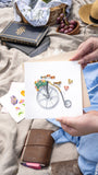Quilled Antique High-Wheel Bicycle being held at picnic next to books, bowl of fruit, and blue dress