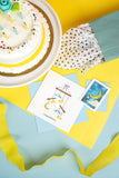 Quilled Baby Clothesline Greeting Card with light blue envelope next to Quilled Moon & Stars Gift Enclosure Mini Card surrounded by gift back and cake on yellow background