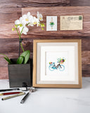 Quilled Bicycle & Flower Basket Greeting Card in gold frame next to white orchid on white marble table in front of wooden wall
