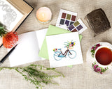 Quilled Bicycle & Flower Basket Greeting Card with light green envelope next to cup of tea, stamps, and book with flower in it