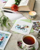 Quilled Bicycle & Flower Basket Greeting Card with light green envelope next to stamps, teacups, and book with flower in it
