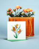 Quilled Bird of Paradise card standing on green envelope sitting in front of bamboo bag filled with yellow flowers in front of light blue background