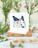 Quilled Birthday Flowers & Blue Butterflies Greeting Card sitting on a book on top of a wooden table, next to pink flower vases and dry flowers.