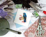 Quilled Bluebird on Flower Branch Greeting Card lays on a white wooden surface, next to gardening gloves, seed packets, a watering can, a cloth, gardening scissors, a gardening shovel, and a pile of dirt.