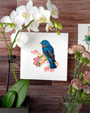 Quilled Bluebird on Flower Branch Greeting Card hung on a wooden wall, next to a white orchid, a vase of pink flowers, and the Quilled Bromeliad Macrame Hanger Gift Enclosure Mini Card.