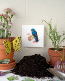 Quilled Bluebird on Flower Branch Greeting Card sitting on a white brick wall, next to pink flowers in terracotta, a ceramic butterfly, packets of flower seeds, a gardening shovel, and a pile of dirt.