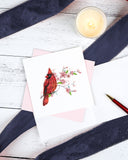 Quilled Cardinal & Cherry Blossom Greeting Card with pink envelope next to open insert with red pen on top of navy ribbon next to candle on top of white wooden table