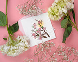 Quilled Cherry Blossoms Greeting Card laying on a pink backdrop, surrounded by flowers.