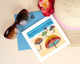 quilled colorful umbrella greeting card with blue envelope next to sunglasses and sunhat on beige background