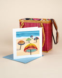 Quilled colorful umbrella greeting card with blue envelope in front of bamboo handbag on beige background