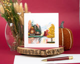 Quilled Cozy Autumn Cabin Greeting Card standing up with dark red envelope behind insert with pen next to pumpkin decor and dried flowers