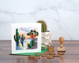 Quilled Desert Landscape Greeting Card standing up in front of a marble background, a cactus plant, above a wooden surface, while next to wax presses and golden wax seals.