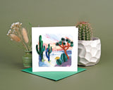 Quilled Desert Landscape Greeting Card standing up in front of an olive green background next to a cactus plant and baby's breath flowers.