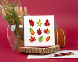 Quilled Fall Foliage Leaves Greeting Card standing on wooden platter on top of insert with pen on red background and pumpkin decor and dried leaves