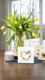 Quilled Floral Heart Wreath Greeting Card sitting on a table in front of a vase of yellow tulips, a candle, and handmade children mother's day cards.