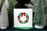 Quilled Holiday Wreath with Ornaments Greeting Card standing on top of green envelope in front of christmas tree decorations