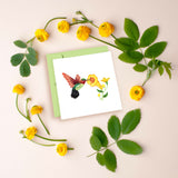 Quilled Hummingbird greeting card laying flat on a cream colored background surrounded by yellow flowers and green leaves arranged in a circle around it. 