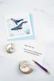 Quilled Humpback Whales Greeting Card with blue envelope on white table next to card insert wishing the recipient a happy hump day with shells and a purple pen