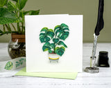 Quilled Monstera greeting card with green envelope in front of plant next to pen quill on green background