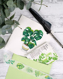 Quilled Monstera greeting card with green envelope next to quilled pen and plant on white wooden background