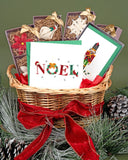 Quilled NOEL Christmas Card sitting in a basket with Quilled Nutcracker Note Card Box Set, Quilled Angel Ornaments Box Set, Quilled Snowflake Ornaments Box Set, Quilled Christmas Ornaments Box Set, and Quilled Poinsettia Ornaments Box Set, surrounded by pine needles and pine cones.